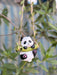 A small, decorative panda figurine hanging from a vine-wrapped rope swing. The panda is holding a green bamboo branch and has pink paw pads, set against a blurred background of green garden leaves.