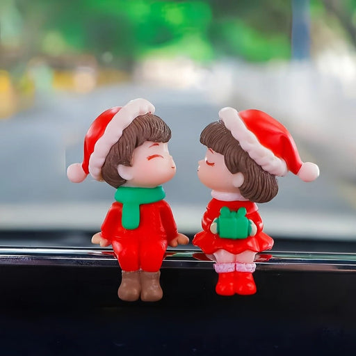 Close-up of two miniature resin figurines, a boy and a girl, dressed in red Santa outfits and hats, sitting side-by-side on the edge of a white textured pot. The boy wears a green scarf; the girl holds a small green present. Ideal for Christmas terrarium or garden decor.