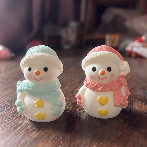 A close-up shot of two small, charming resin snowman figurines sitting on a wooden surface. The snowman on the left wears a pink hat and scarf, and the one on the right wears a light blue hat and scarf. Both have small black eyes, a carrot nose, and a cheerful smile.