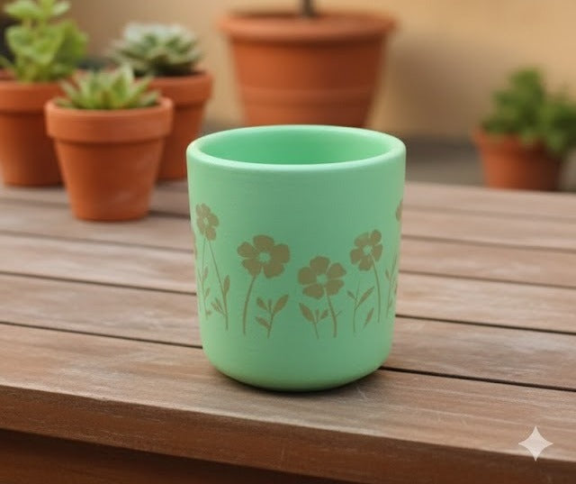 A small, round, light mint green terracotta planter sitting on a wooden plank surface. The pot has a golden-brown silhouette design of wild flowers.