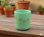 A small, round, light mint green terracotta planter sitting on a wooden plank surface. The pot has a golden-brown silhouette design of wild flowers.