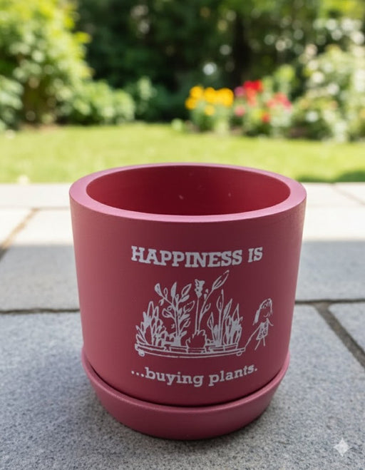Close-up of a round, vibrant brown terracotta planter and saucer sitting on grey patio stones. The pot is decorated with a white graphic illustration of plants and the text "HAPPINESS IS ...buying plants." A blurred green garden is visible in the background
