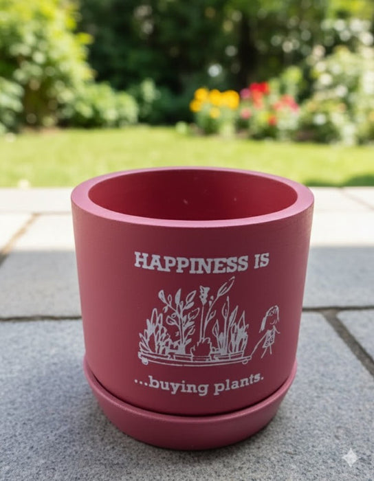 Close-up of a round, vibrant brown terracotta planter and saucer sitting on grey patio stones. The pot is decorated with a white graphic illustration of plants and the text "HAPPINESS IS ...buying plants." A blurred green garden is visible in the background