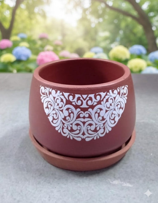 A round, rich coffee-brown terracotta planter and matching saucer sitting on a grey surface. The pot has a detailed white scroll design on the front. The background is a blurred, sunny garden of colourful flowers.