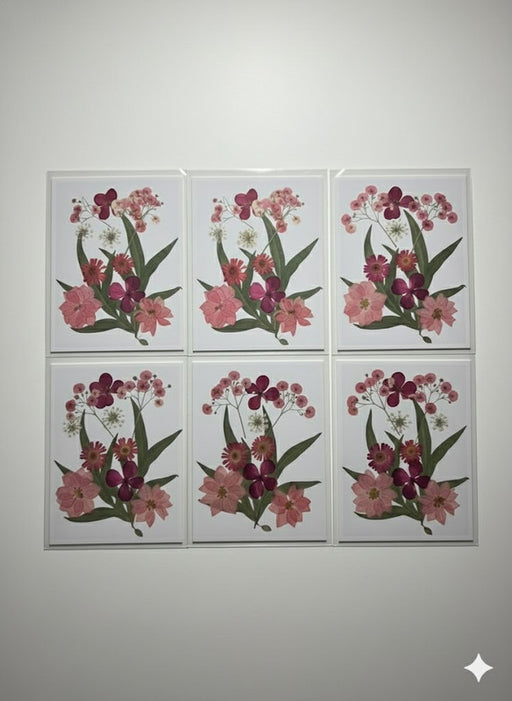 A close-up image of a handcrafted art piece featuring a bouquet arrangement of real, pressed pink and red flowers, including zinnias and baby's breath, sealed on a white background.