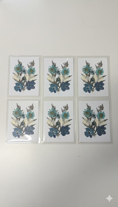Set of six white cards displayed on a light wooden floor, each featuring an intricate arrangement of blue, teal, and white pressed flowers, including daisies and hydrangeas.