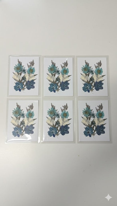 Set of six white cards displayed on a light wooden floor, each featuring an intricate arrangement of blue, teal, and white pressed flowers, including daisies and hydrangeas.