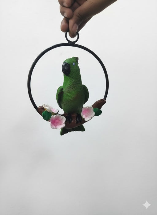 A hand holding a circular black metal ring with a small, textured green parrot figurine perched on a brown branch decorated with pink and white flowers.