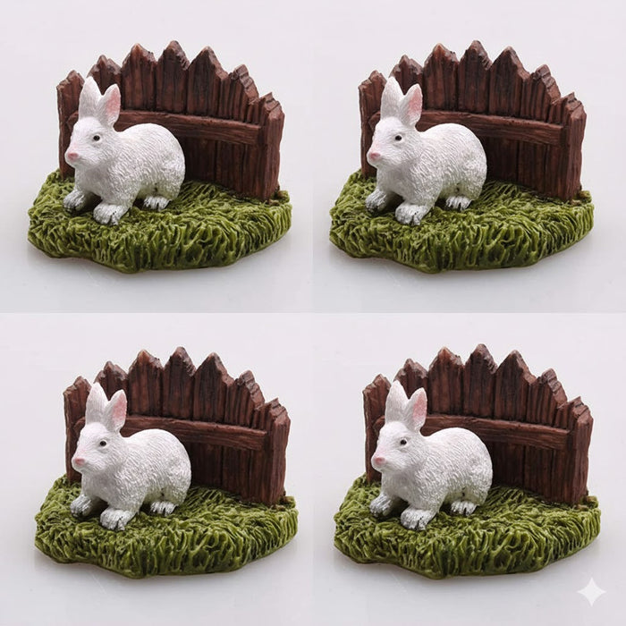 Four white miniature resin rabbit figurines sitting on green grass in front of a small brown wooden fence.