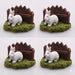 Four white miniature resin rabbit figurines sitting on green grass in front of a small brown wooden fence.