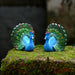 Two bright blue miniature peacock figurines with fully displayed green and gold tails 