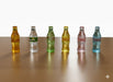 Six miniature glass soda bottles (Coca-Cola and Sprite) lined up on a wooden surface, featuring different colors: amber Coke, clear Sprite, green Coke, yellow Coke, pink Coke, and blue Coke.