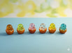 Six tiny, colorful glass or resin chick figurines (brown, green, pink, clear/white, yellow, and blue) emerging from small, orange eggshell-shaped bases, sitting on a light blue surface with a soft, bright yellow and white