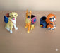 Three colorful resin dog figurines sitting side-by-side on a wooden surface. From left to right: a yellow Labrador Retriever, an orange German Shepherd, and a smaller, fluffy orange dog, each wearing a small backpack that functions as a pen holder or planter.