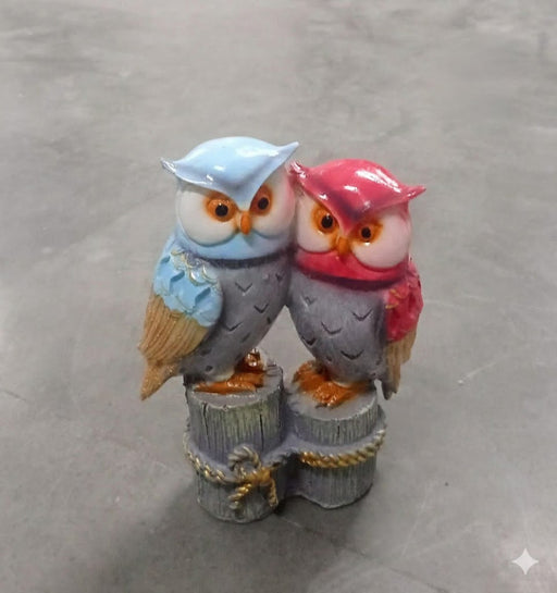 A resin figurine of two colorful owls, one blue and one pink/red, standing together on small wooden posts wrapped with rope.