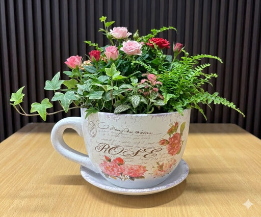 A white ceramic oversized teacup and saucer planter featuring pink rose illustrations, vintage cursive script, and a distressed antique texture on a wooden surface.