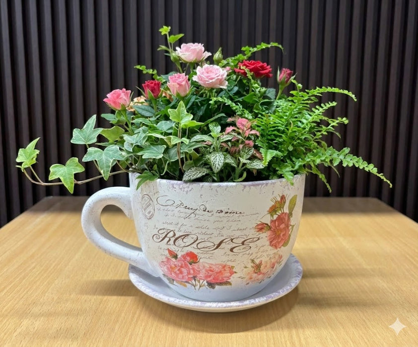A white ceramic oversized teacup and saucer planter featuring pink rose illustrations, vintage cursive script, and a distressed antique texture on a wooden surface.