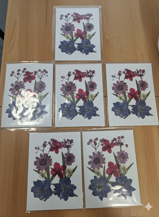 Set of 6 handmade greeting cards, each featuring an arrangement of real pressed flowers in shades of deep blue, purple, and magenta, sealed in individual plastic sleeves and displayed on a wooden surface.