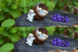Set of two identical images showing tiny ceramic white bunny rabbit figurines, one leaning against a small brown woven pot containing two other bunnies, next to a cluster of purple flowers, all resting on a dark wood slice outdoors.
