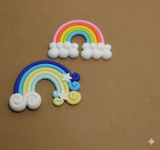 Two colorful, stylized rainbow cabochons or appliqués made of clay or resin, featuring white clouds, stars, and swirls, set against a plain brown background.