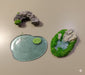 Three miniature terrain pieces: a gray rocky arch or cave entrance, a flat piece of resin simulating a small pond with a lily pad, and an oval base with a miniature pond, moss, and a small rock feature.