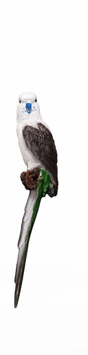 A close-up, full-body photograph of a parakeet figurine, or garden statue, perched on a branch. The bird has a white head, a blue cere above its beak, dark gray and white wings, and a long green and gray tail.
