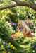 Cute ceramic or resin squirrel/chipmunk garden ornament swinging from a tree branch on a rope swing in a lush, sunny garden with colorful flowers in the background. (
