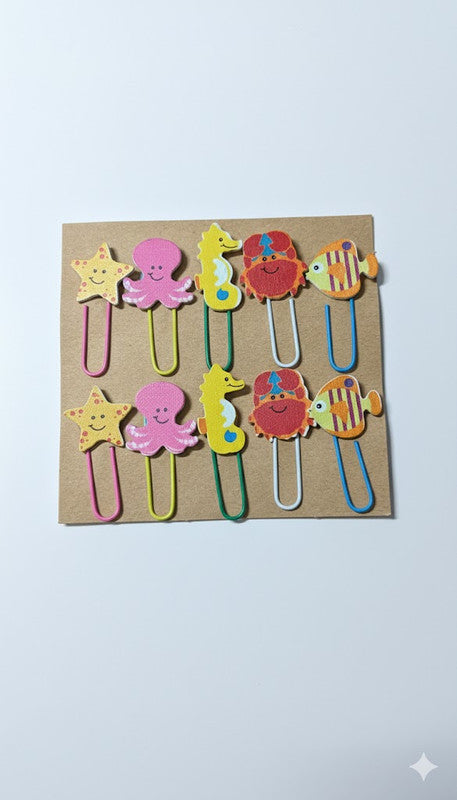 A set of ten colorful wooden sea life paper clips, arranged on a brown card background. Designs include a yellow starfish, pink octopus, yellow seahorse, red crab/lobster, and striped fish, with assorted colored metal clips.