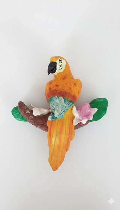 A small, brightly colored ceramic or clay figurine of an orange and yellow macaw or parrot perched on a brown branch with green leaves and pink and white tropical flowers.