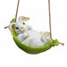 A small white resin bunny figurine with yellow accents, relaxing on its back in a vibrant green fabric hammock swing suspended by rope.