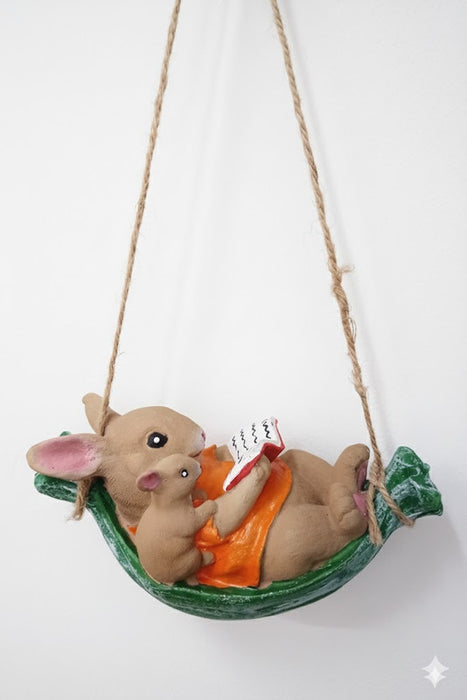 Decorative figurine of a bunny and a mouse reading a book together while relaxing in a green hammock, hanging by twine rope against a white wall.