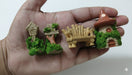 A set of four miniature fairy garden accessories held in a hand, including a wooden birdhouse, a rustic moss-covered wooden chair, a small wooden bench, and a whimsical stone mushroom house covered in green moss.