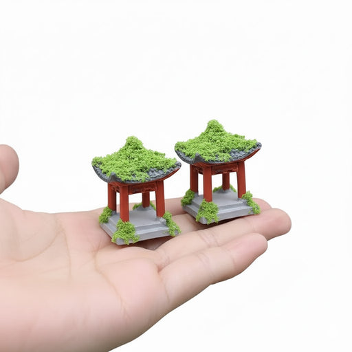"Pair of miniature Chinese-style garden pavilions or pagodas with red pillars, grey base, and green flocking moss on the roof and around the base, held in the palm of a hand."