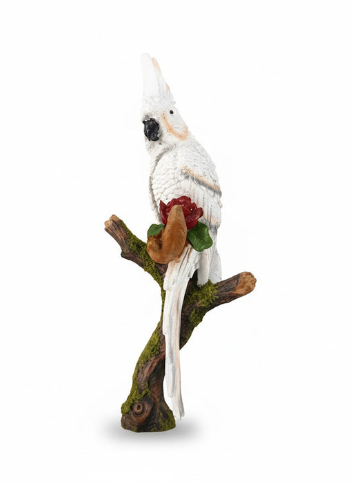 A white Cockatoo bird figurine with its crest raised, perched on a moss-covered tree branch, holding a single red rose in one of its claws.