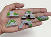 Miniature terrain pieces featuring grey rocky formations with bright green moss held in a hand. The set includes small hills, a stepped cave entrance, and a rock archway, ideal for tabletop gaming or wargaming.
