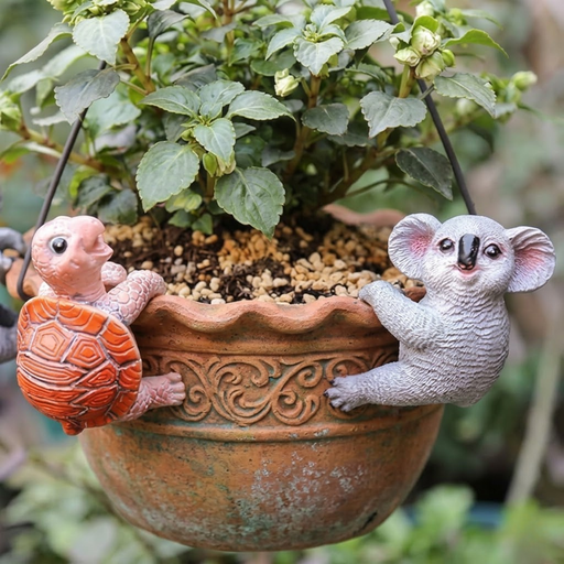 Two small resin animal figurines, a bright orange turtle and a grey koala, hanging from the rim of a black outdoor flower pot with green leafy plants.