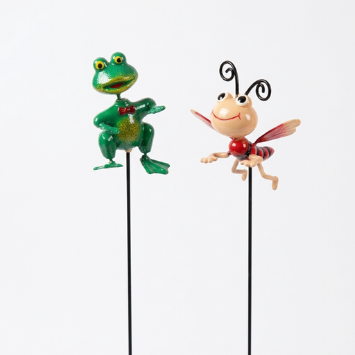 Two whimsical metal garden stakes featuring a glittery green frog and a smiling cartoon ladybug attached to thin black rods, displayed against a white wire grid and wood panel background.