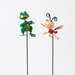 Two whimsical metal garden stakes featuring a glittery green frog and a smiling cartoon ladybug attached to thin black rods, displayed against a white wire grid and wood panel background.