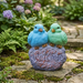 A colorful outdoor garden statue featuring two plump songbirds—one sky blue and one mint green—perched side-by-side on a rustic, purple-gray stone-textured base embossed with the word "Welcome" in a friendly script.