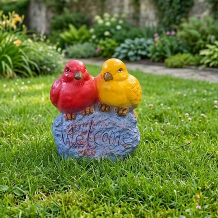 A vibrant garden welcome statue featuring a pair of colorful red and yellow lovebirds perched on a grey, stone-textured base with the word "Welcome" carved into it, set on a green artificial lawn.