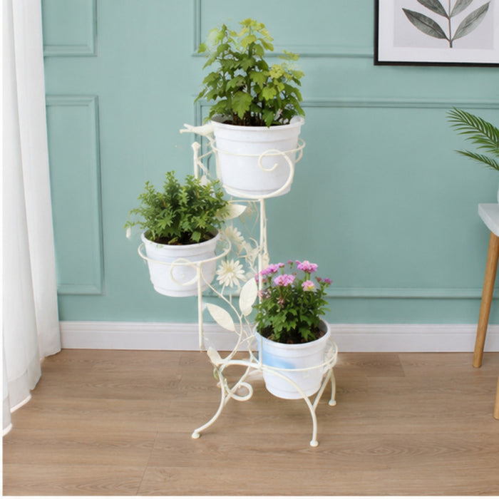 Cream-white decorative metal plant stand featuring floral vines, leaf cutouts, and bird accents, holding a white potted plant against a textured wall.