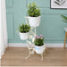 Cream-white decorative metal plant stand featuring floral vines, leaf cutouts, and bird accents, holding a white potted plant against a textured wall.