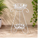 White metal two-tier plant stand with decorative leaf patterns and scrollwork holding a potted green plant against a textured wall.