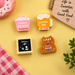 Set of four cute food and drink miniatures arranged on a yellow background, including a pink "Sweet Dessert" cupcake, a yellow "Drink Me" beer mug, a coffee-themed sign, and an amber "Cake" piece, alongside a red-and-white checkered cloth and a wooden sign that reads "Life is Sweeter with Good Food".