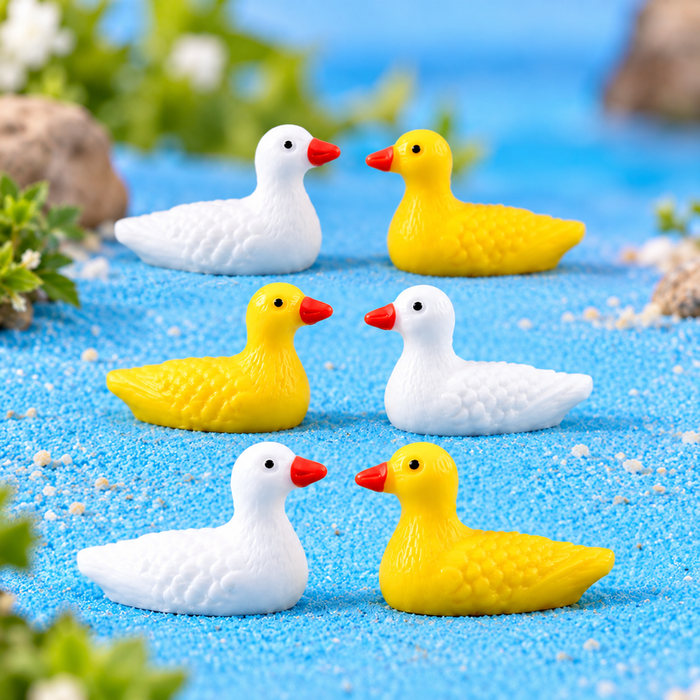 Two miniature resin ducks (one white and one bright yellow) placed in a fairy garden micro landscape with blue sand water and green grass.