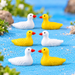 Two miniature resin ducks (one white and one bright yellow) placed in a fairy garden micro landscape with blue sand water and green grass.