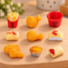 A 9-piece set of miniature KFC-themed toy food items—including fried chicken, french fries, a bucket, a tart, and nuggets—arranged on a wooden kitchen countertop.