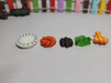 A set of five miniature items lined up on a white surface, including a small blue-and-white patterned plate, an orange textured food item, dark clustered pieces, green lettuce, and a small cooked meat piece.