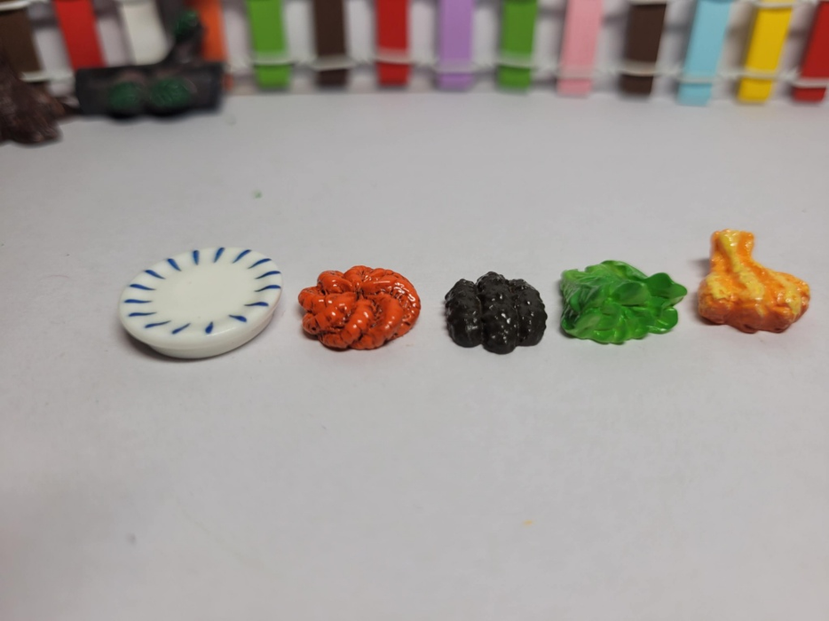 A set of five miniature items lined up on a white surface, including a small blue-and-white patterned plate, an orange textured food item, dark clustered pieces, green lettuce, and a small cooked meat piece.