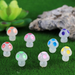 A set of 8 colorful miniature mushroom figurines with translucent stems and vibrant polka-dot caps arranged on green artificial moss next to a succulent and decorative rock.